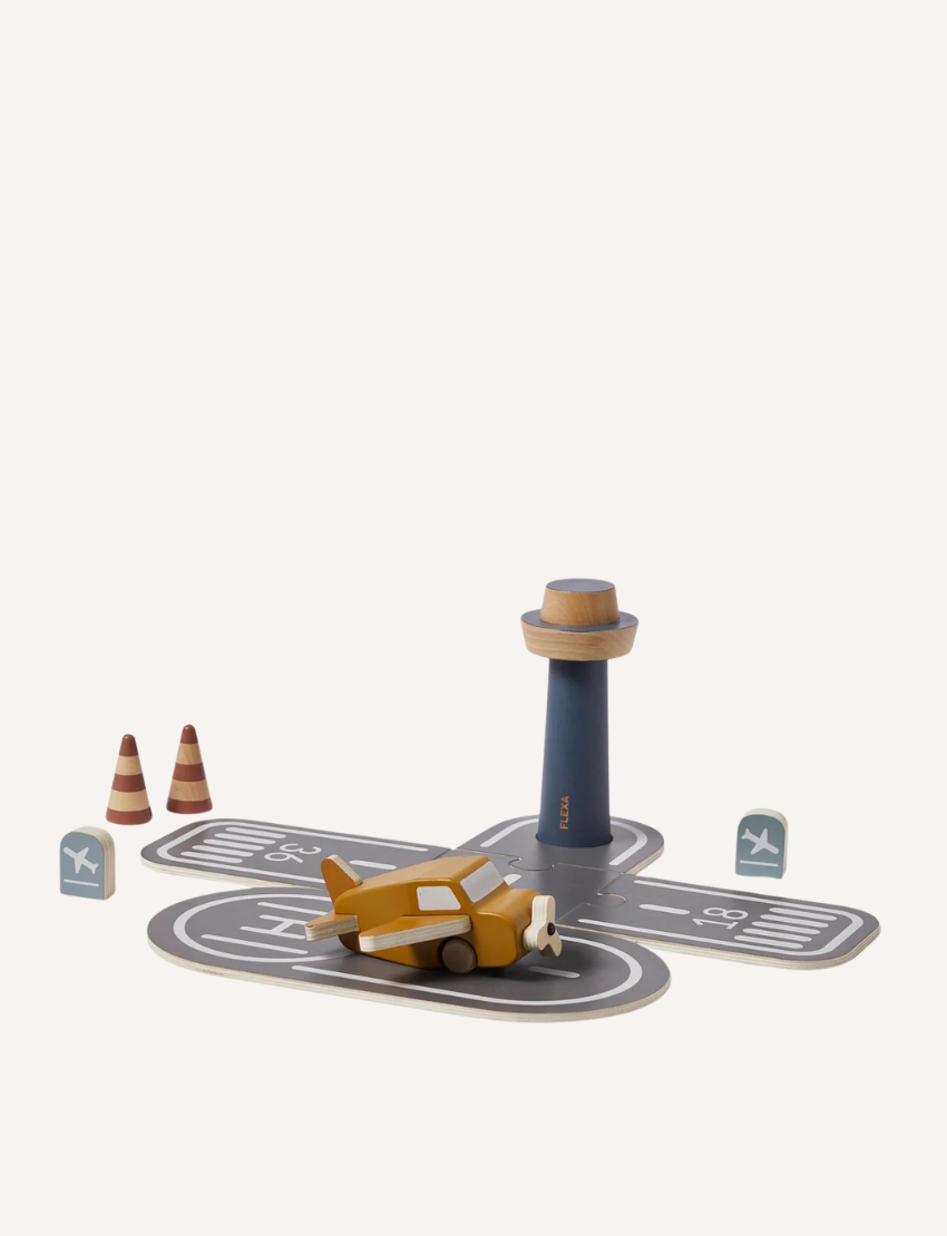 A small toy airplane from the Flexa Wooden Airport collection is positioned on a grey runway play mat featuring white markings. Nearby, you can find a cylindrical control tower, two orange traffic cones, and two teal airport signs. All items are displayed against a plain white background.