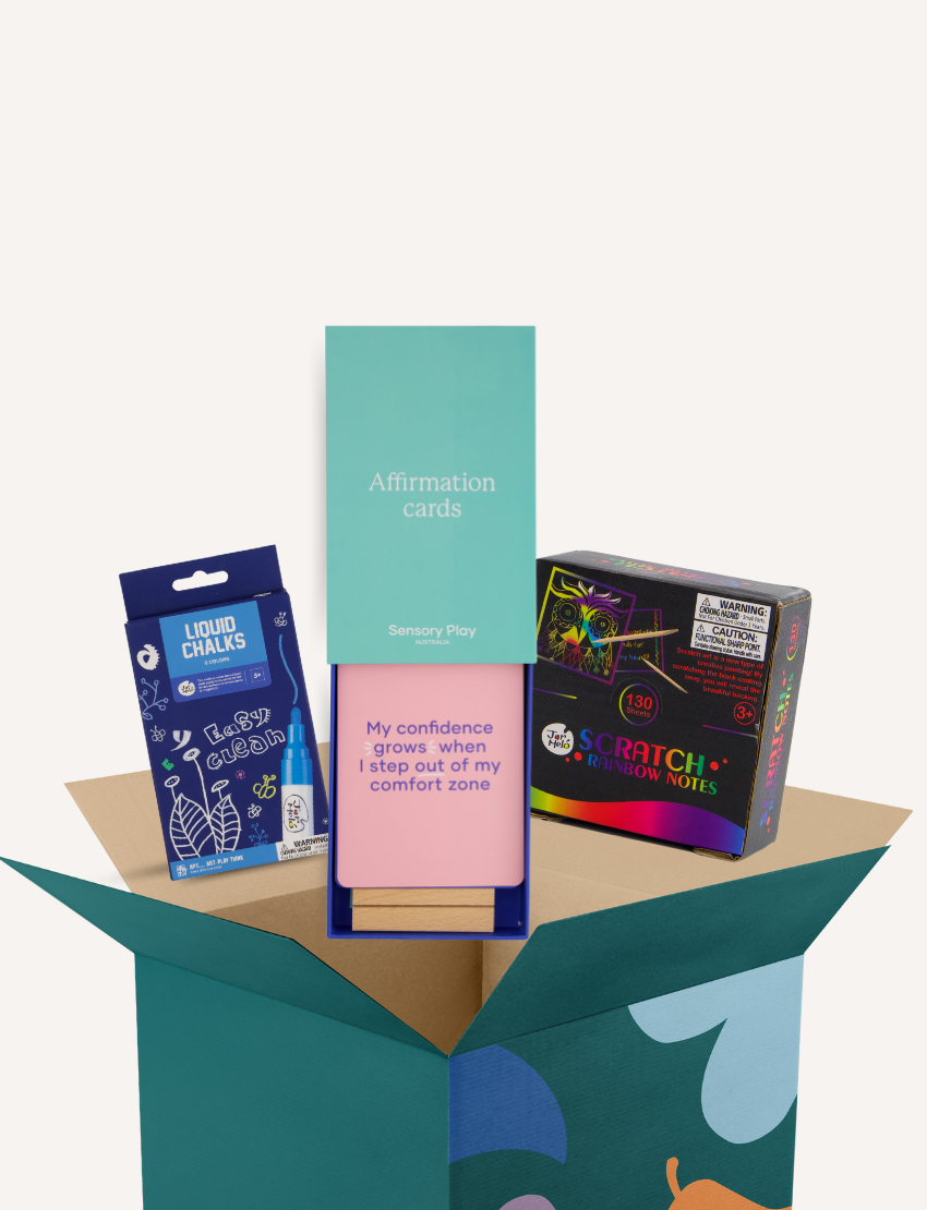 A cardboard box from The Play Way's Mindset Kit contains a package of Liquid Chalks, a pack of Sensory Play Affirmation Cards, and a set of Scratch Rainbow Notes. The Affirmation Cards' package is prominently displayed, with the message reading, "My confidence grows when I step out of my comfort zone.