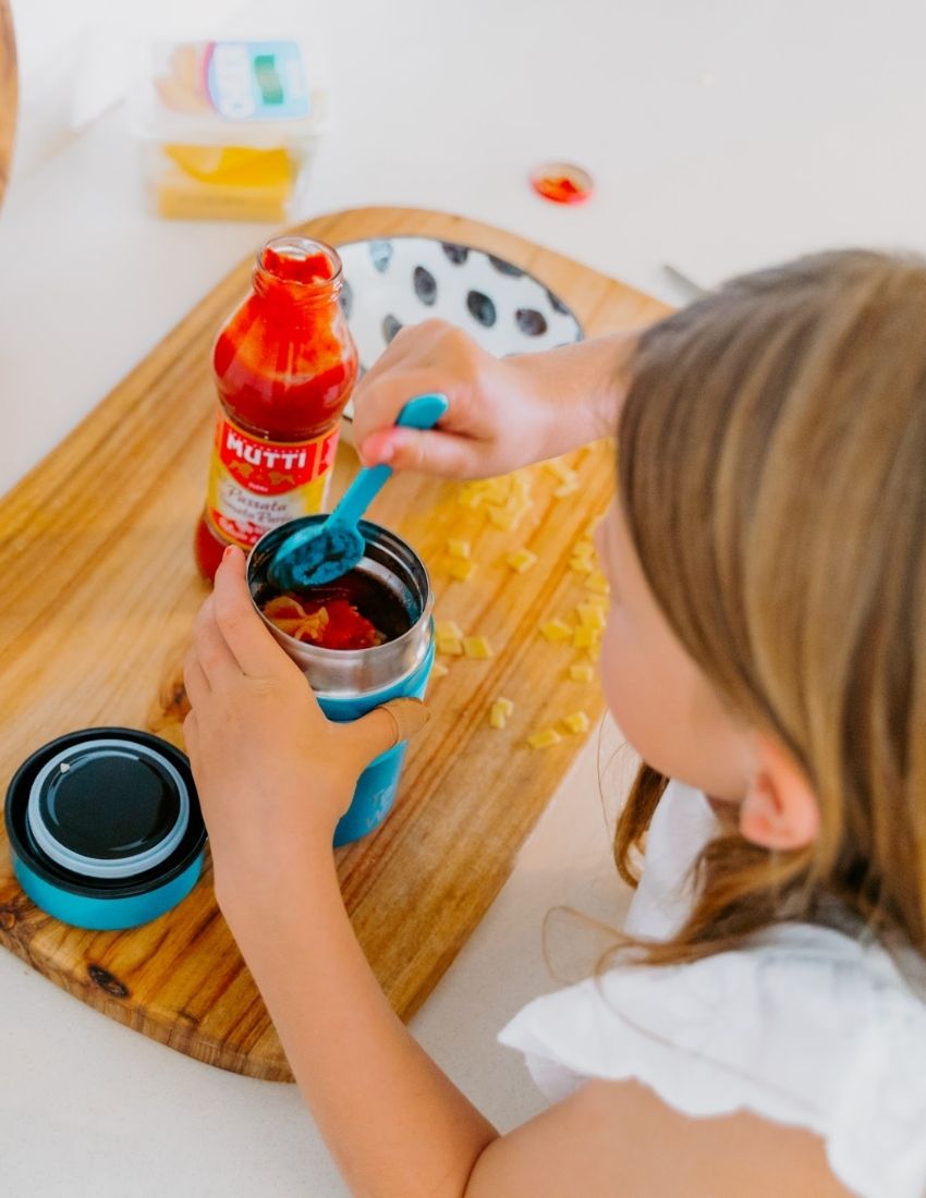 A blue insulated food jar from The Play Way with the initials "T P W" printed on the front. The Lunch Buddy Insulated Container Set features a matching blue silicone band around its midsection that holds a blue spoon, along with a leak-proof lid. The background is plain white.