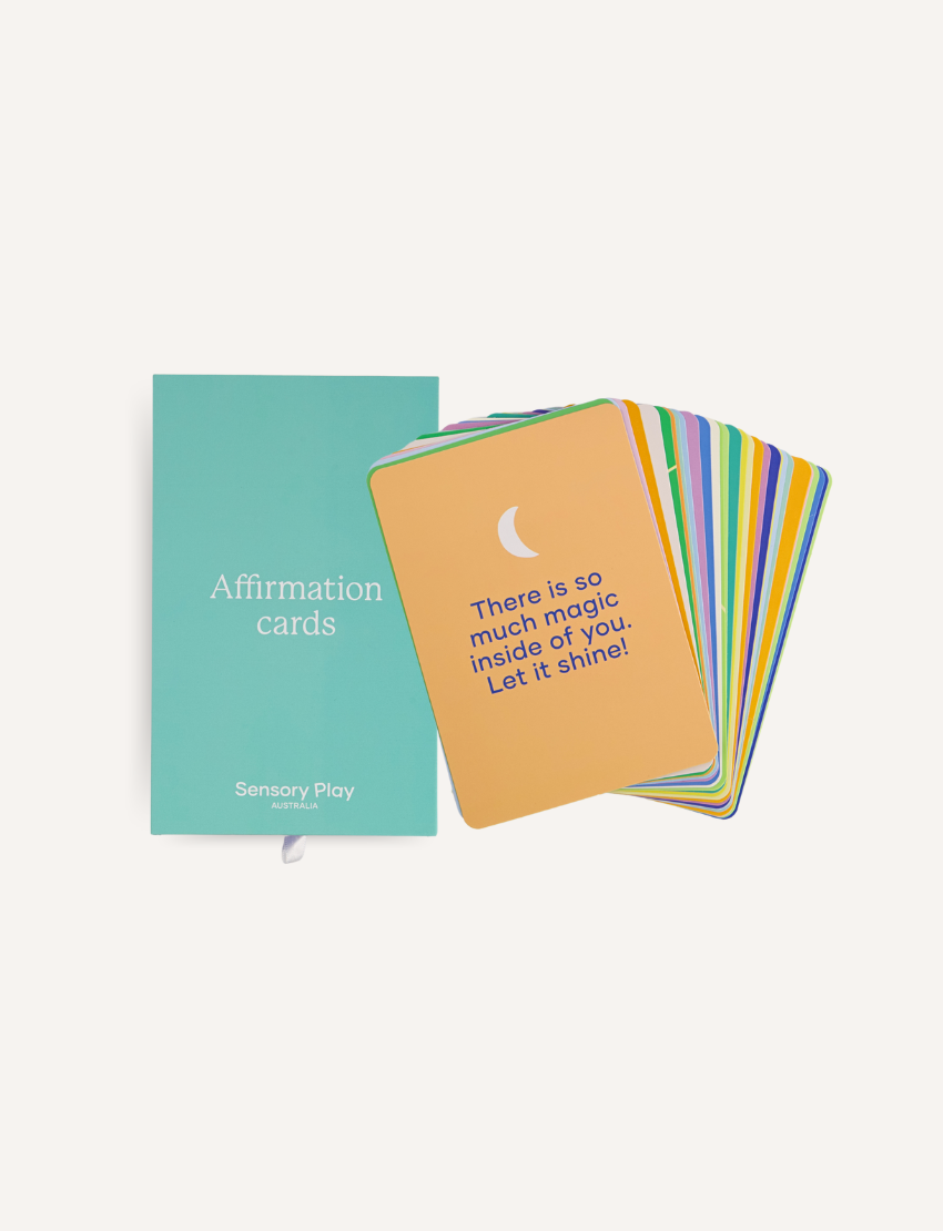 A cardboard box from The Play Way's Mindset Kit contains a package of Liquid Chalks, a pack of Sensory Play Affirmation Cards, and a set of Scratch Rainbow Notes. The Affirmation Cards' package is prominently displayed, with the message reading, "My confidence grows when I step out of my comfort zone.