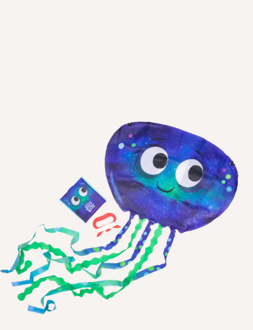 A young girl in a purple sweater and black pants holds a Jelly Kite by Tiger Tribe, designed as a blue jellyfish with big eyes and adorned with green and blue streamers. She is standing on a grassy field with trees in the background.