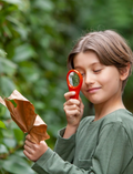 A child is seated at a table with an open notebook from Tiger Tribe's Outdoor Activity Set. The child is arranging leaves and small shells on a page. Nearby, there is a box with a hand resting on it, and the child is wearing a striped shirt.