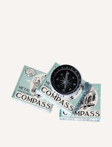 A metal compass with a black face is placed on top of two boxes. The boxes are labeled "Adventurer's Compass" by House of Marbles and display illustrations and text on a light blue background. The compass needle is pointing north.