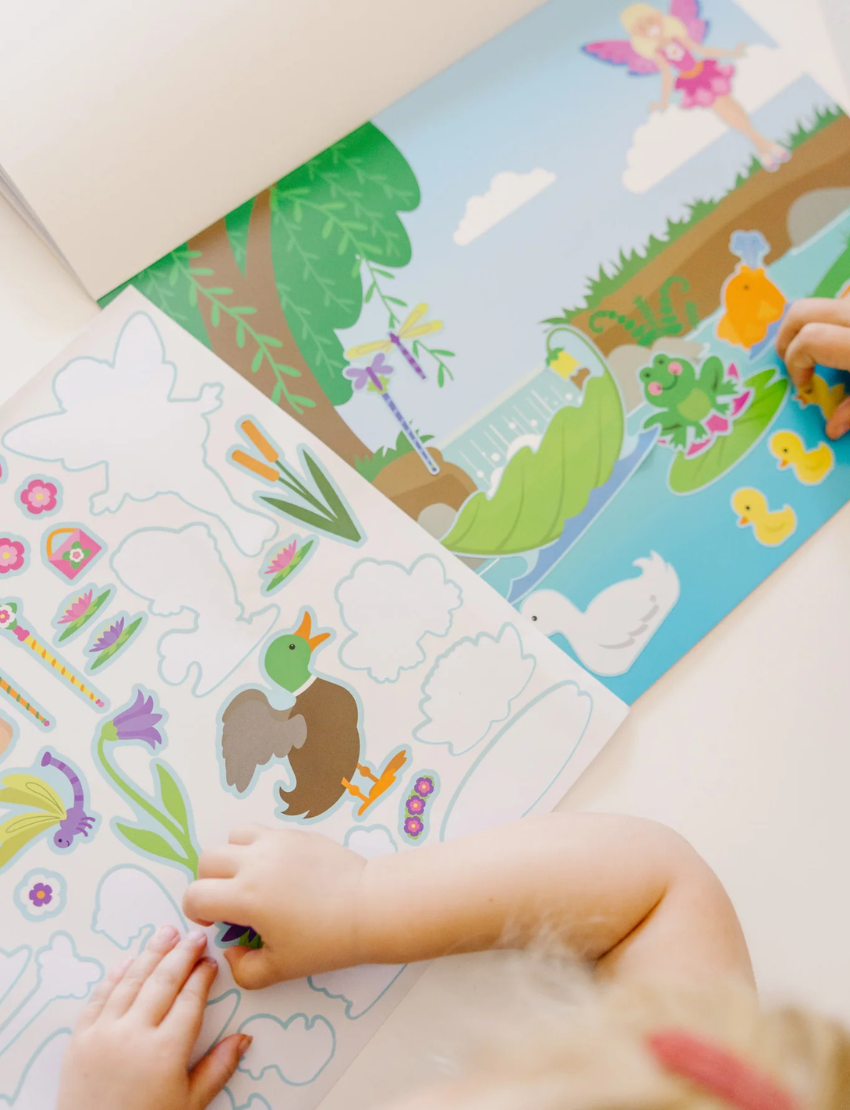 A child plays with a Melissa and Doug Reusable Sticker Pad - Fairies, which features a vibrant pond scene. The page includes stickers of a fairy, frog, duck, ducklings, swan, and various plants. The child is in the process of peeling off a duck sticker from the colorful and playful scene.