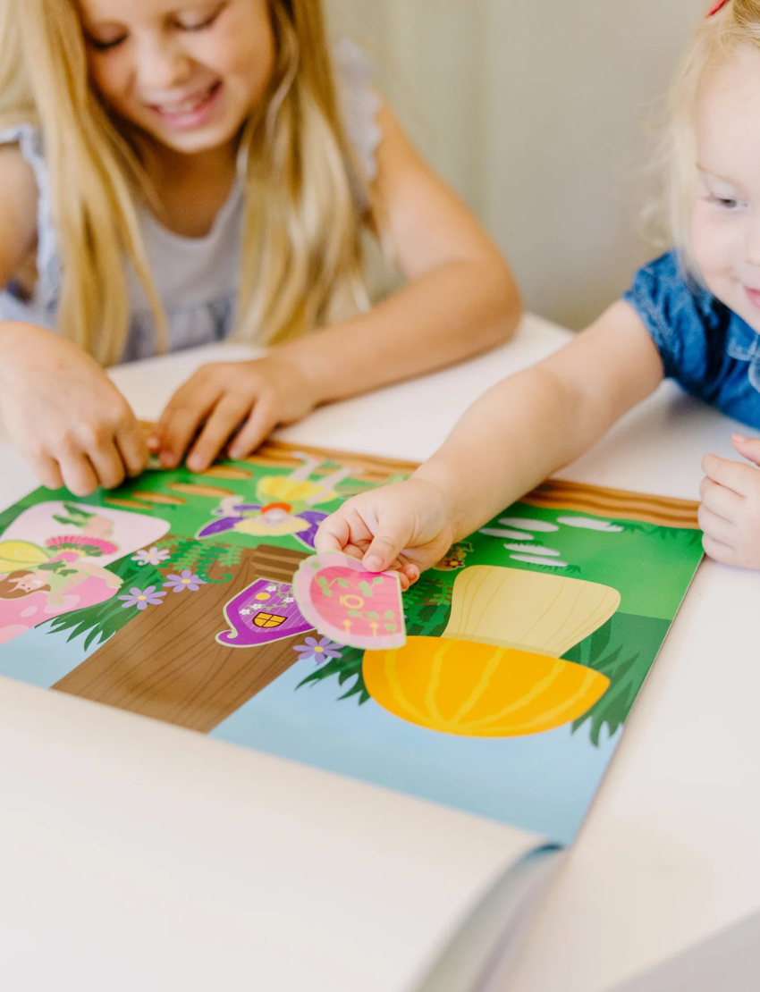 A child plays with a Melissa and Doug Reusable Sticker Pad - Fairies, which features a vibrant pond scene. The page includes stickers of a fairy, frog, duck, ducklings, swan, and various plants. The child is in the process of peeling off a duck sticker from the colorful and playful scene.