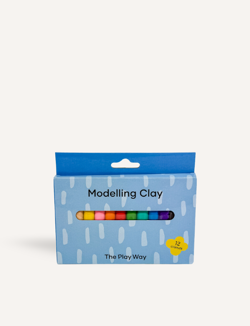 A vibrant rainbow crafted from The Play Way Modelling Clay rests on a wooden table. Beside it sits a blue box labeled "The Play Way Modelling Clay." In the background, there's a plant in a pot adorned with a yellow and white checkered design.