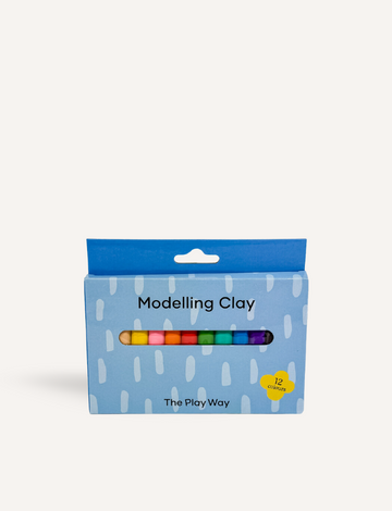 A vibrant rainbow crafted from The Play Way Modelling Clay rests on a wooden table. Beside it sits a blue box labeled "The Play Way Modelling Clay." In the background, there's a plant in a pot adorned with a yellow and white checkered design.