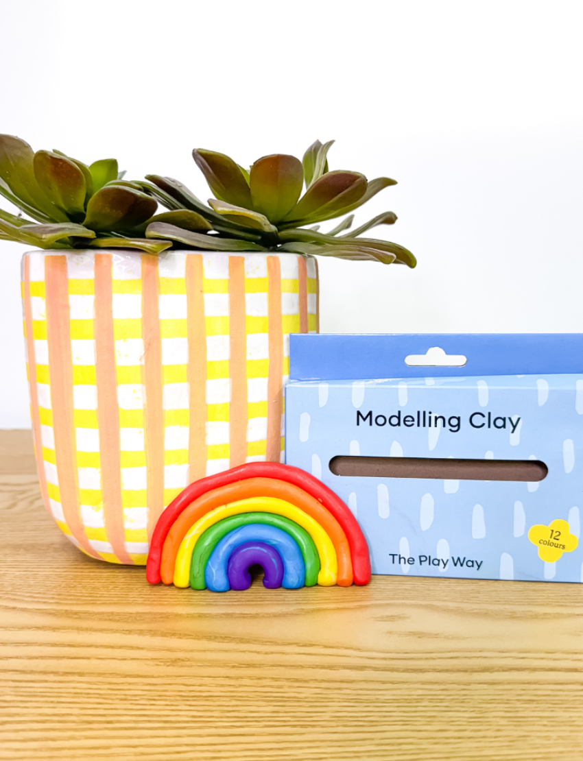 A vibrant rainbow crafted from The Play Way Modelling Clay rests on a wooden table. Beside it sits a blue box labeled "The Play Way Modelling Clay." In the background, there's a plant in a pot adorned with a yellow and white checkered design.