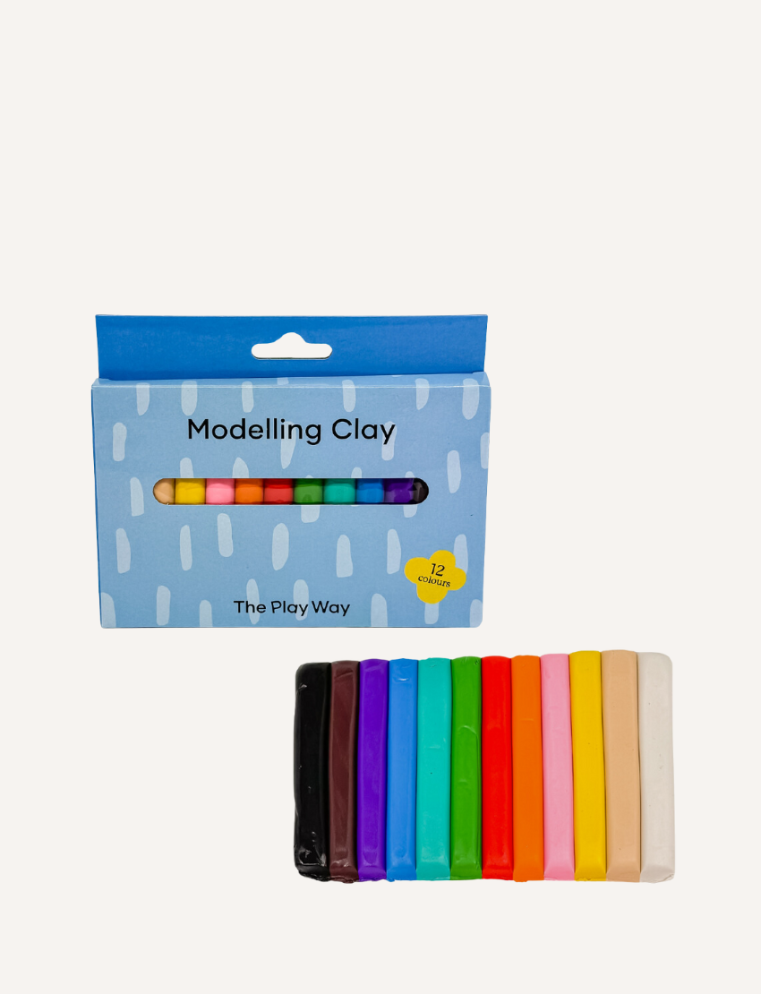 A vibrant rainbow crafted from The Play Way Modelling Clay rests on a wooden table. Beside it sits a blue box labeled "The Play Way Modelling Clay." In the background, there's a plant in a pot adorned with a yellow and white checkered design.