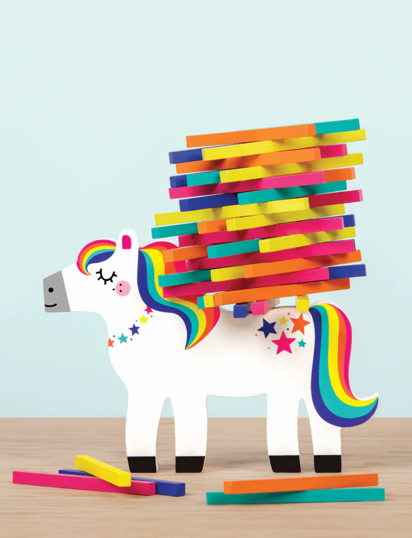 A vibrant box of the "Pony Pile Up Balancing Game" by Mudpuppy, showcasing an illustration of a white pony with a rainbow mane and tail. The pony's back is piled with colorful sticks. The text on the box reads "Stack & Balance To Win" and "Wooden Family Fun Balancing Game.