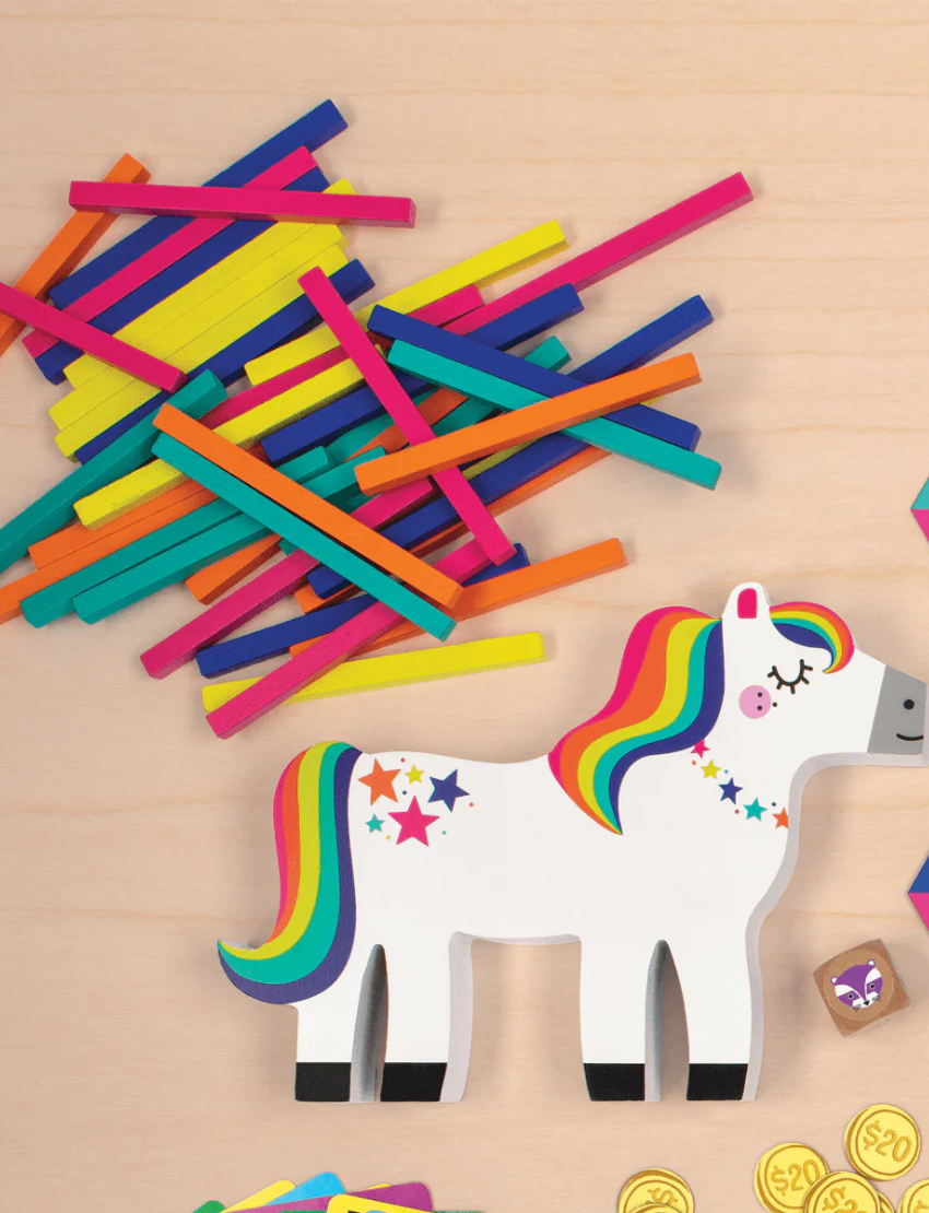 A vibrant box of the "Pony Pile Up Balancing Game" by Mudpuppy, showcasing an illustration of a white pony with a rainbow mane and tail. The pony's back is piled with colorful sticks. The text on the box reads "Stack & Balance To Win" and "Wooden Family Fun Balancing Game.