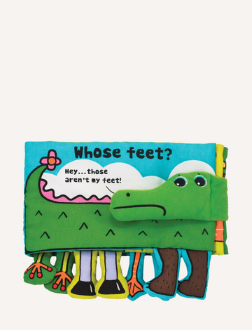 The "Whose Feet? Soft Book" by Melissa and Doug is a vibrant children's fabric book showcasing an illustration of a green creature, possibly a crocodile or dinosaur, with playful eyes. One page features different animal legs alongside a speech bubble declaring, "Hey... those aren't my feet!