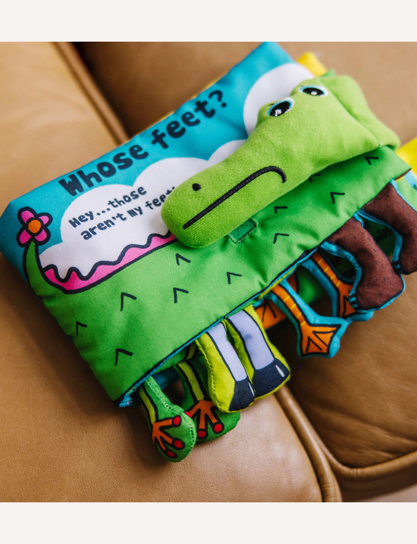 The "Whose Feet? Soft Book" by Melissa and Doug is a vibrant children's fabric book showcasing an illustration of a green creature, possibly a crocodile or dinosaur, with playful eyes. One page features different animal legs alongside a speech bubble declaring, "Hey... those aren't my feet!