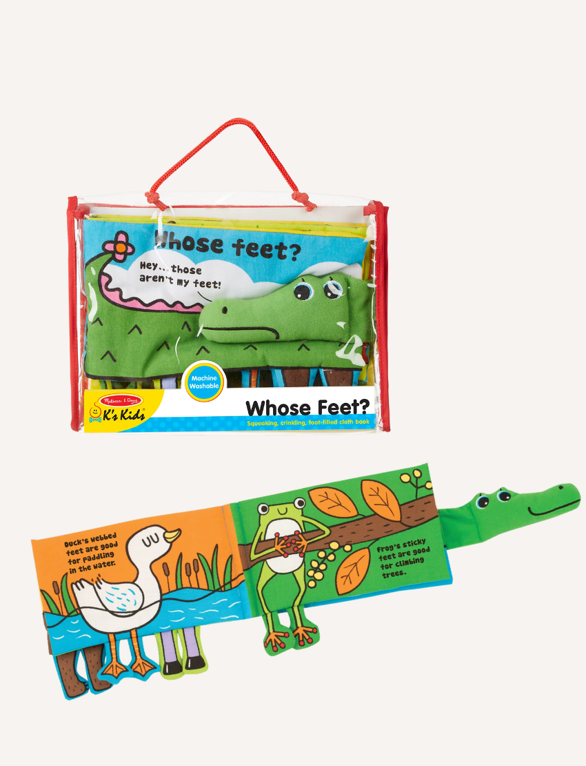 The "Whose Feet? Soft Book" by Melissa and Doug is a vibrant children's fabric book showcasing an illustration of a green creature, possibly a crocodile or dinosaur, with playful eyes. One page features different animal legs alongside a speech bubble declaring, "Hey... those aren't my feet!