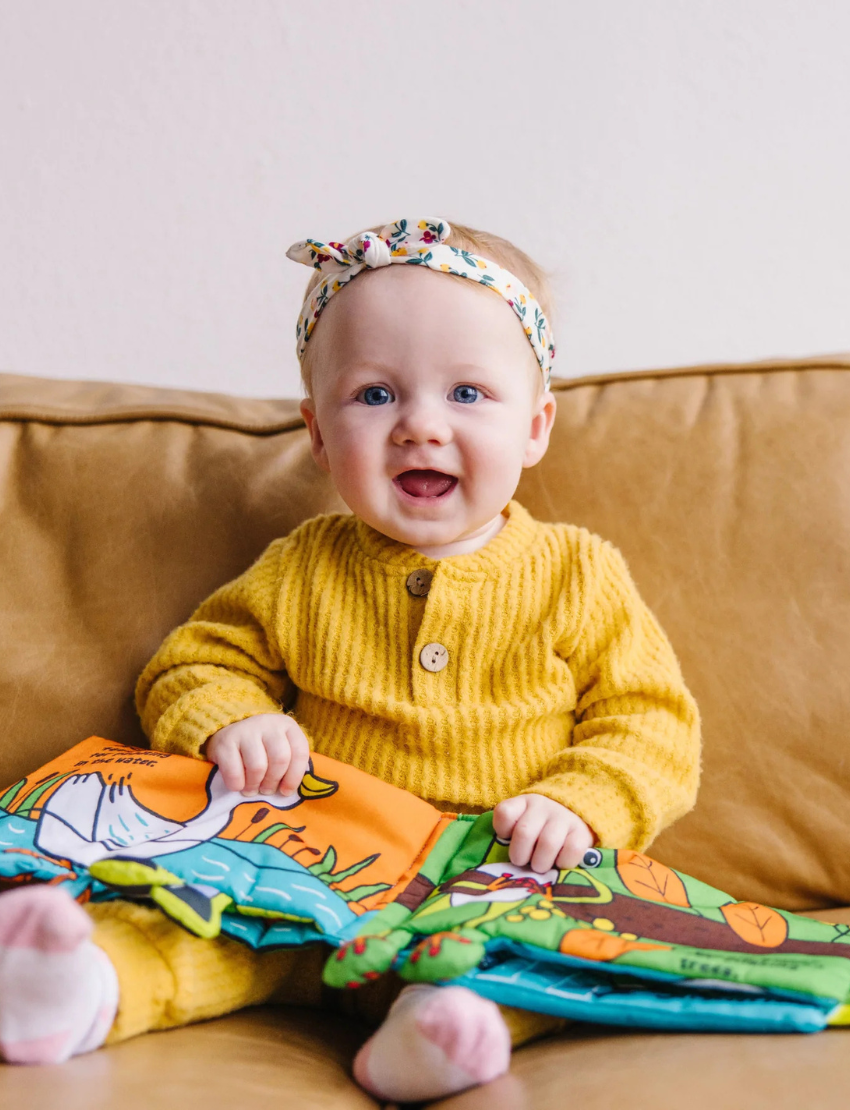 The "Whose Feet? Soft Book" by Melissa and Doug is a vibrant children's fabric book showcasing an illustration of a green creature, possibly a crocodile or dinosaur, with playful eyes. One page features different animal legs alongside a speech bubble declaring, "Hey... those aren't my feet!