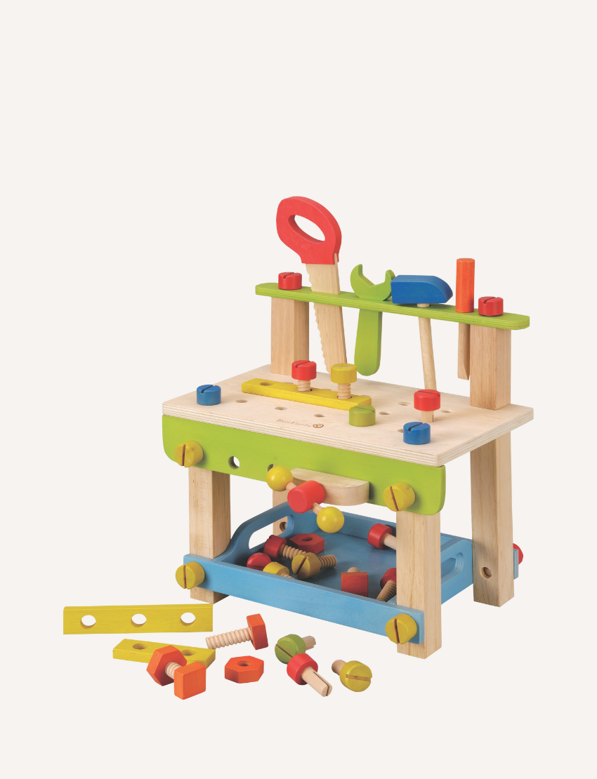 An eye-catching **Play Large Work Bench with Tools** from **Ever Earth** is positioned on a table. This vibrant wooden toy tool bench comes complete with various tools, including a wrench, hammer, screwdriver, and bolts, all showcased in vivid hues of red, blue, green, and yellow. The background reveals a room adorned with large windows.