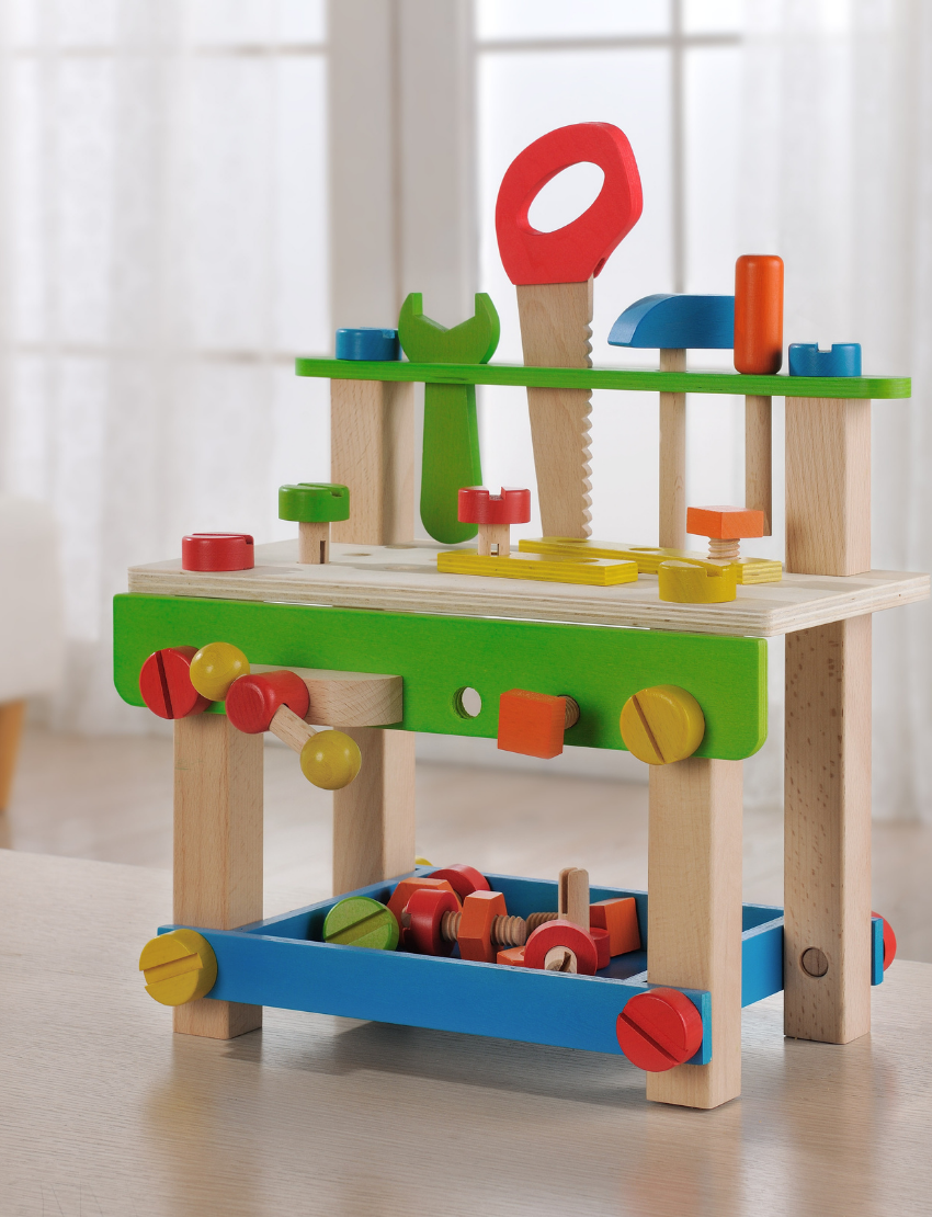 An eye-catching **Play Large Work Bench with Tools** from **Ever Earth** is positioned on a table. This vibrant wooden toy tool bench comes complete with various tools, including a wrench, hammer, screwdriver, and bolts, all showcased in vivid hues of red, blue, green, and yellow. The background reveals a room adorned with large windows.