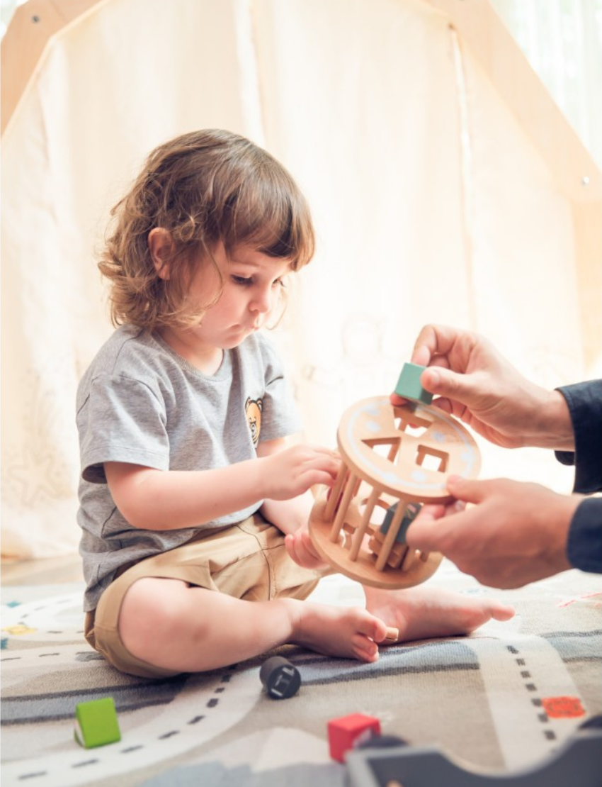 A toddler with curly hair, dressed in a grey T-shirt and khaki shorts, sits on a patterned mat playing with the Ever Earth Pull Along Recycling Truck - Pastel. An adult hand is helping place a green block into the truck. In the background, soft furnishings can be seen.