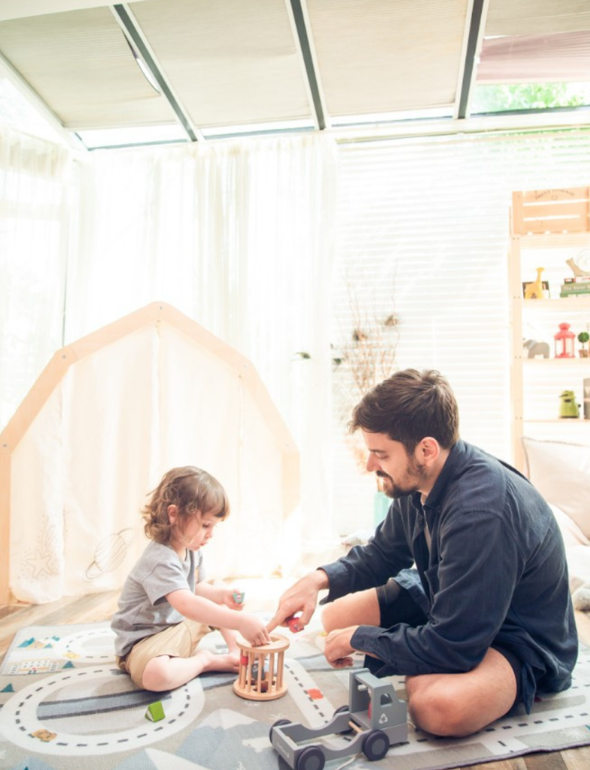 A toddler with curly hair, dressed in a grey T-shirt and khaki shorts, sits on a patterned mat playing with the Ever Earth Pull Along Recycling Truck - Pastel. An adult hand is helping place a green block into the truck. In the background, soft furnishings can be seen.