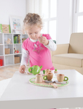 A young girl wearing a pink top with white sleeves is standing next to a white table, holding a small spoon. The table features the Ever Earth Tea Set, including a green teapot and several wooden tea cups. In the background, there is a shelf with books and toys and a beige couch.