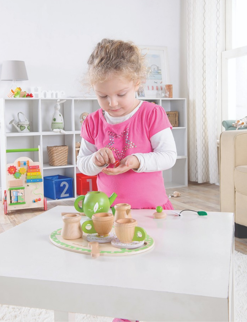 A young girl wearing a pink top with white sleeves is standing next to a white table, holding a small spoon. The table features the Ever Earth Tea Set, including a green teapot and several wooden tea cups. In the background, there is a shelf with books and toys and a beige couch.