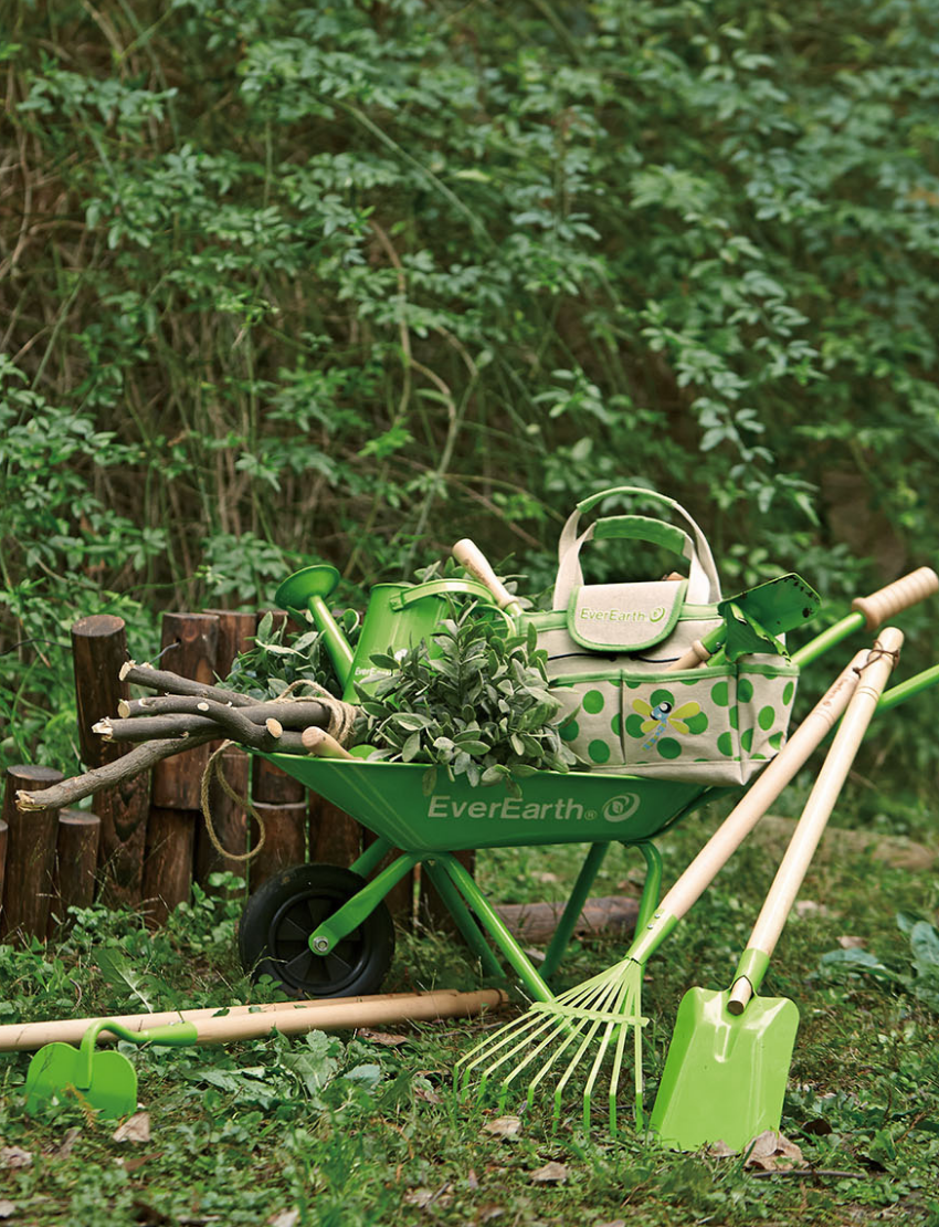 An "Ever Earth" green wheelbarrow filled with branches and foliage sits on grass next to gardening tools, including a rake, shovel, and hand trowel. An Outdoor Gardening Bag With Tools in a beige and green polka-dotted design rests on top of the wheelbarrow. In the background, there are green shrubs and a wooden fence—perfect for outdoor play.