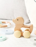 A brown cardboard box contains an Ever Earth Babies Gift Set (In a Gift Box), making it ideal as a baby shower gift. The box features a small clear window on the front, showcasing the wooden toys inside, and is adorned with illustrations of ducks and flowers. Labeled 