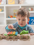 The Educational Pull-along Musical Crocodile by Ever Earth is a wooden pull-along toy train featuring colorful elements. The front section displays a frog face, followed by a segment adorned with rainbow-colored xylophone bars, and the back resembles a bear holding a bell. Connected by string and rolling on wooden wheels, this charming toy provides both fun and learning for young children.