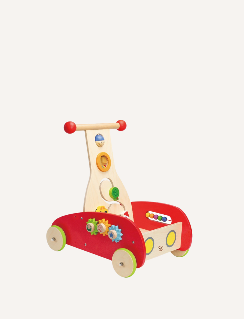 A toddler wearing a light blue shirt and shorts pushes the Hape Wonder Walker, a red and wooden toy. The front panel of the Wonder Walker is adorned with colorful beads and features, against a plain white background.