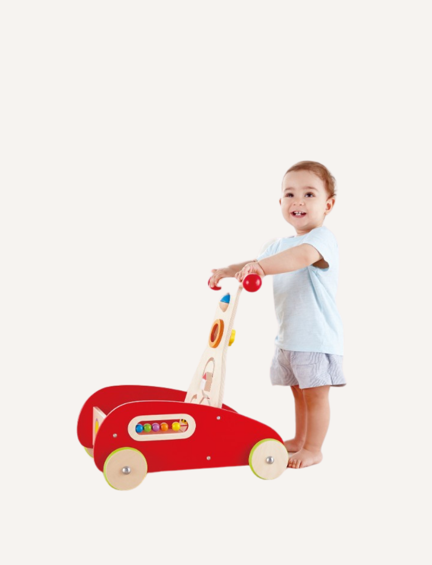 A toddler wearing a light blue shirt and shorts pushes the Hape Wonder Walker, a red and wooden toy. The front panel of the Wonder Walker is adorned with colorful beads and features, against a plain white background.