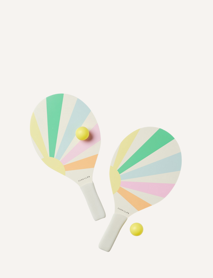 A pair of colorful, pastel rainbow-striped beach paddles with white handles, accompanied by two yellow balls, all neatly stored in a transparent carrying case with a blue zipper. This set is the Pool Side Beach Paddle Set Pastel Gelato by Sunnylife.