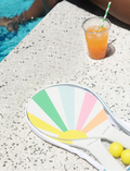 A pair of colorful, pastel rainbow-striped beach paddles with white handles, accompanied by two yellow balls, all neatly stored in a transparent carrying case with a blue zipper. This set is the Pool Side Beach Paddle Set Pastel Gelato by Sunnylife.