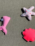 The Melody the Mermaid Dive Buddies Neon Strawberry Set of 3 by Sunnylife features three adorable plush toys on a white background, including a pink mermaid tail, an orange shell, and a white starfish with a smiley face and blue eyes—ideal companions for children's water play.