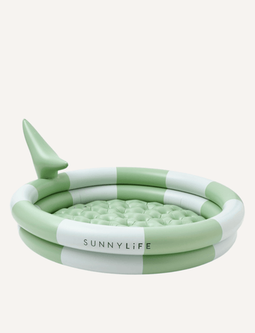 Two children are playing in a small "Shark Tribe Khaki" inflatable backyard pool by Sunnylife in a sunny yard. One child stands in the durable PVC pool and bends over, while the other child, outside the pool, holds a white bucket. The backyard features a larger rectangular swimming pool.