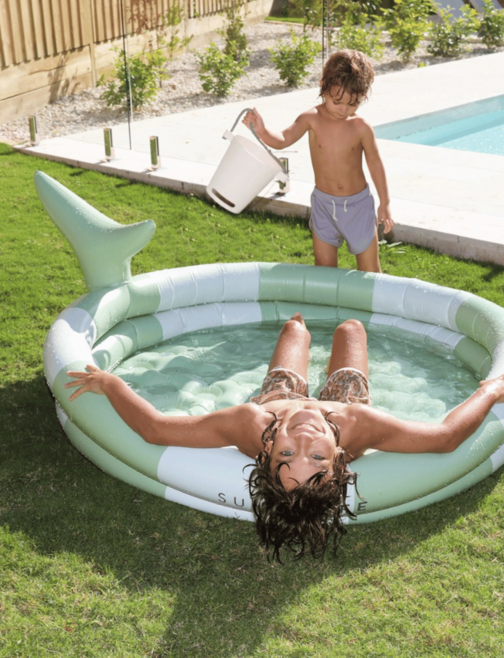 Two children are playing in a small "Shark Tribe Khaki" inflatable backyard pool by Sunnylife in a sunny yard. One child stands in the durable PVC pool and bends over, while the other child, outside the pool, holds a white bucket. The backyard features a larger rectangular swimming pool.