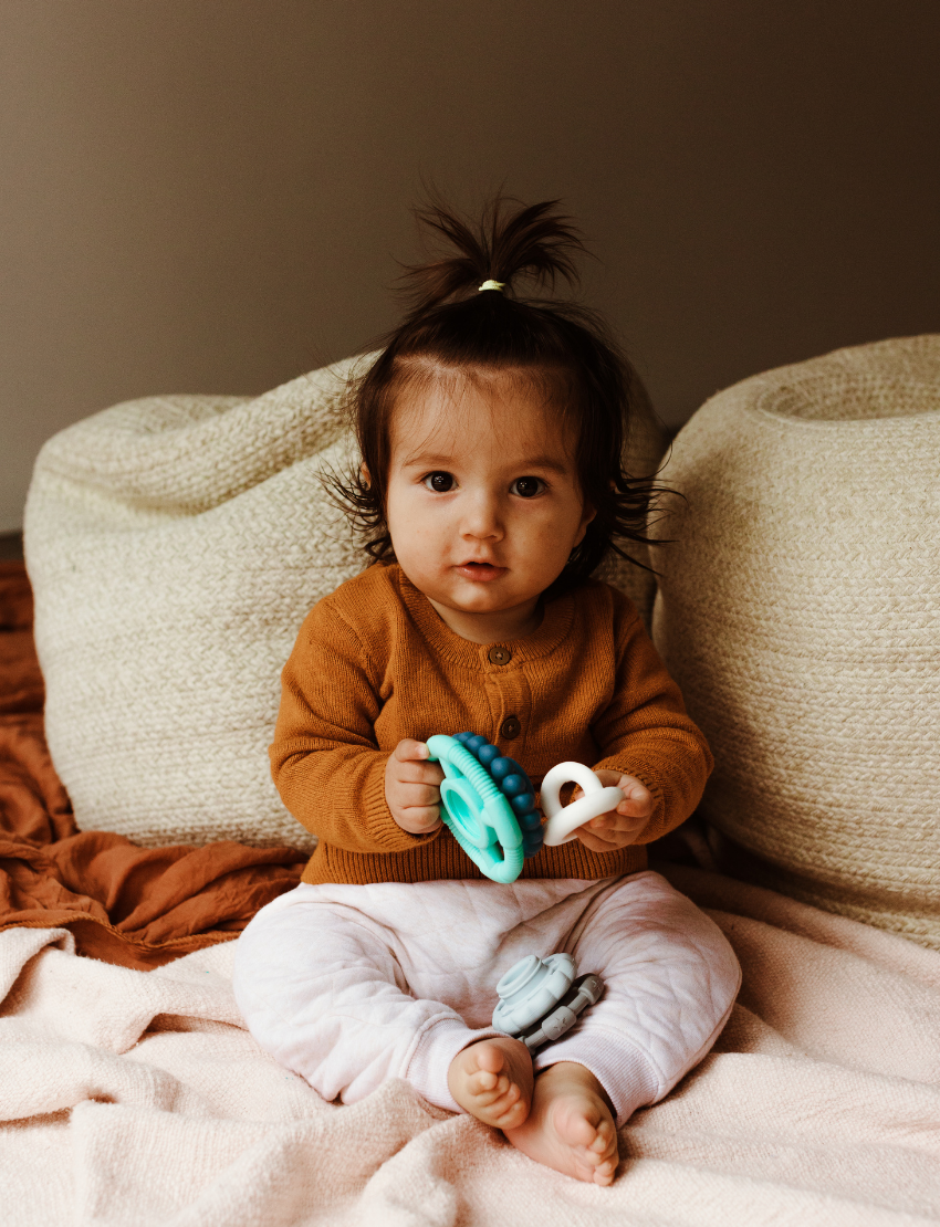 The Rainbow Stacker and Teether Toy by Jellystone Designs features a set of five silicone teething toys in assorted shapes and colors, including two circular ones with textured edges, a flat design, one shaped like a gear, and a smooth white piece. They are neatly arranged on a simple white backdrop.