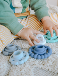 The Rainbow Stacker and Teether Toy by Jellystone Designs features a set of five silicone teething toys in assorted shapes and colors, including two circular ones with textured edges, a flat design, one shaped like a gear, and a smooth white piece. They are neatly arranged on a simple white backdrop.