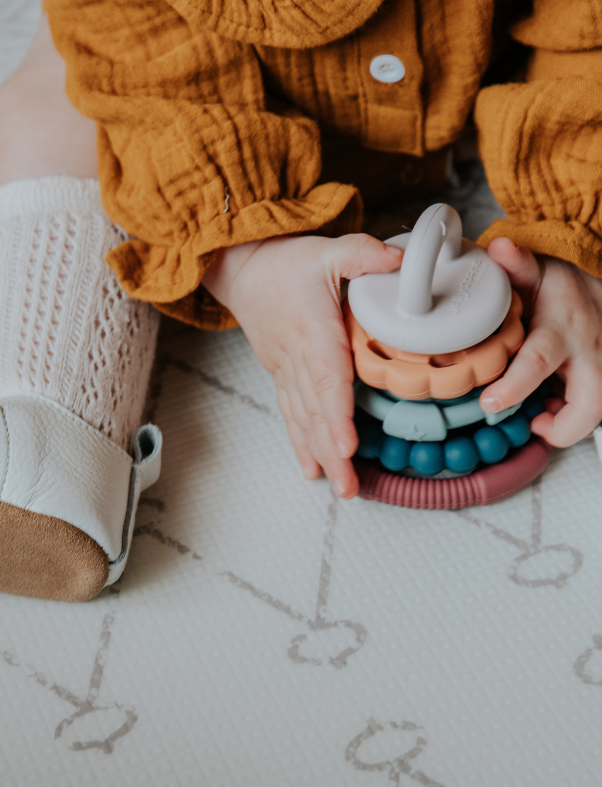 The Rainbow Stacker and Teether Toy by Jellystone Designs features a set of five silicone teething toys in assorted shapes and colors, including two circular ones with textured edges, a flat design, one shaped like a gear, and a smooth white piece. They are neatly arranged on a simple white backdrop.