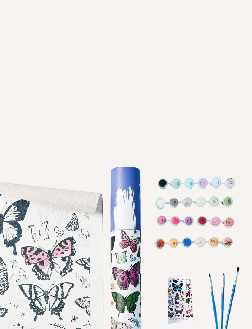 The Journey of Something Paint by Numbers kit includes a rolled canvas featuring butterfly outlines, housed in a blue cylindrical container. The kit also contains numbered paint pots, three paintbrushes, and a reference sheet. The canvas showcases butterflies partially colored in pink and blue hues.