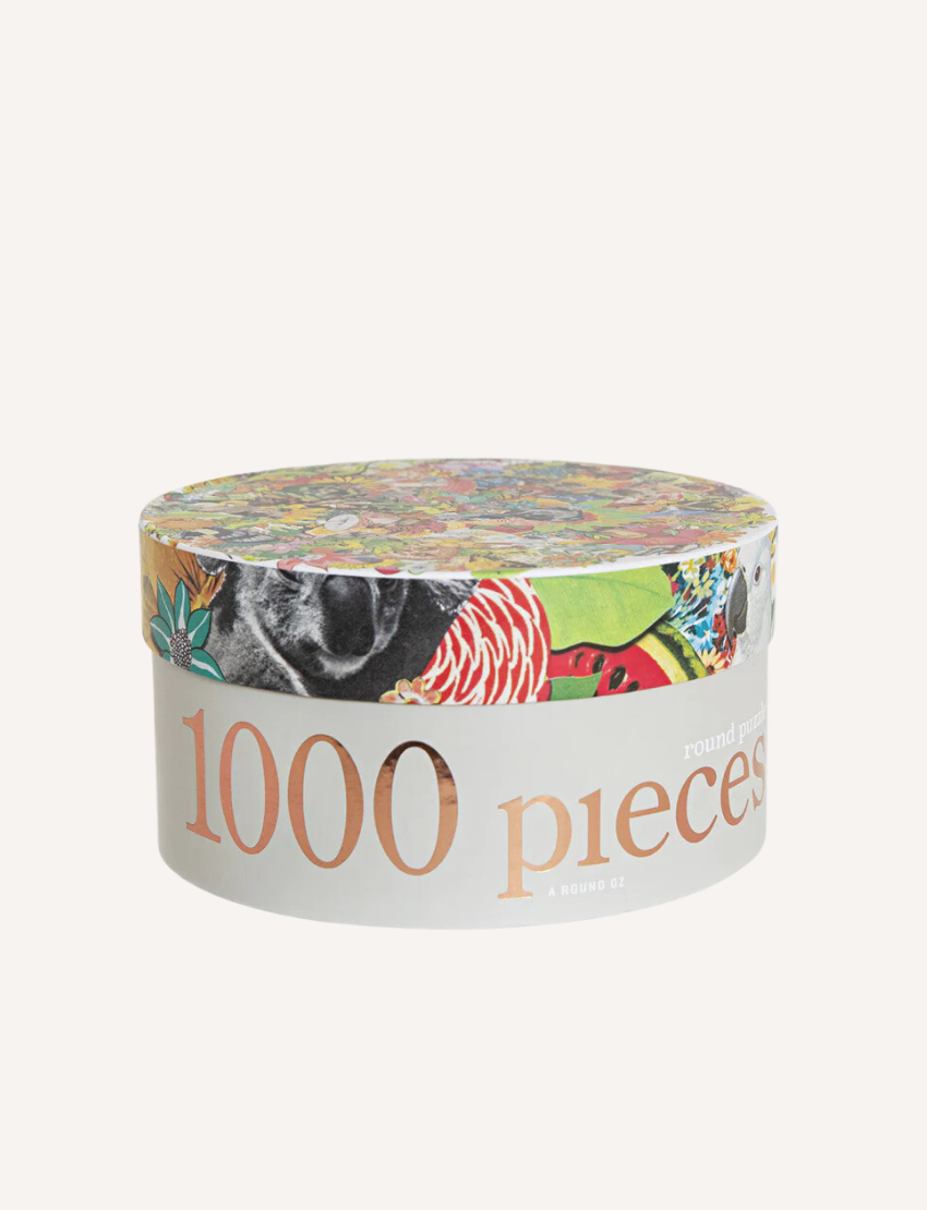 A round, colorful jigsaw puzzle with completed sections and scattered pieces is shown next to an open cylindrical container labeled "1000 Piece Puzzle" by Journey of Something. The puzzle features a vibrant design with various small images and shapes.