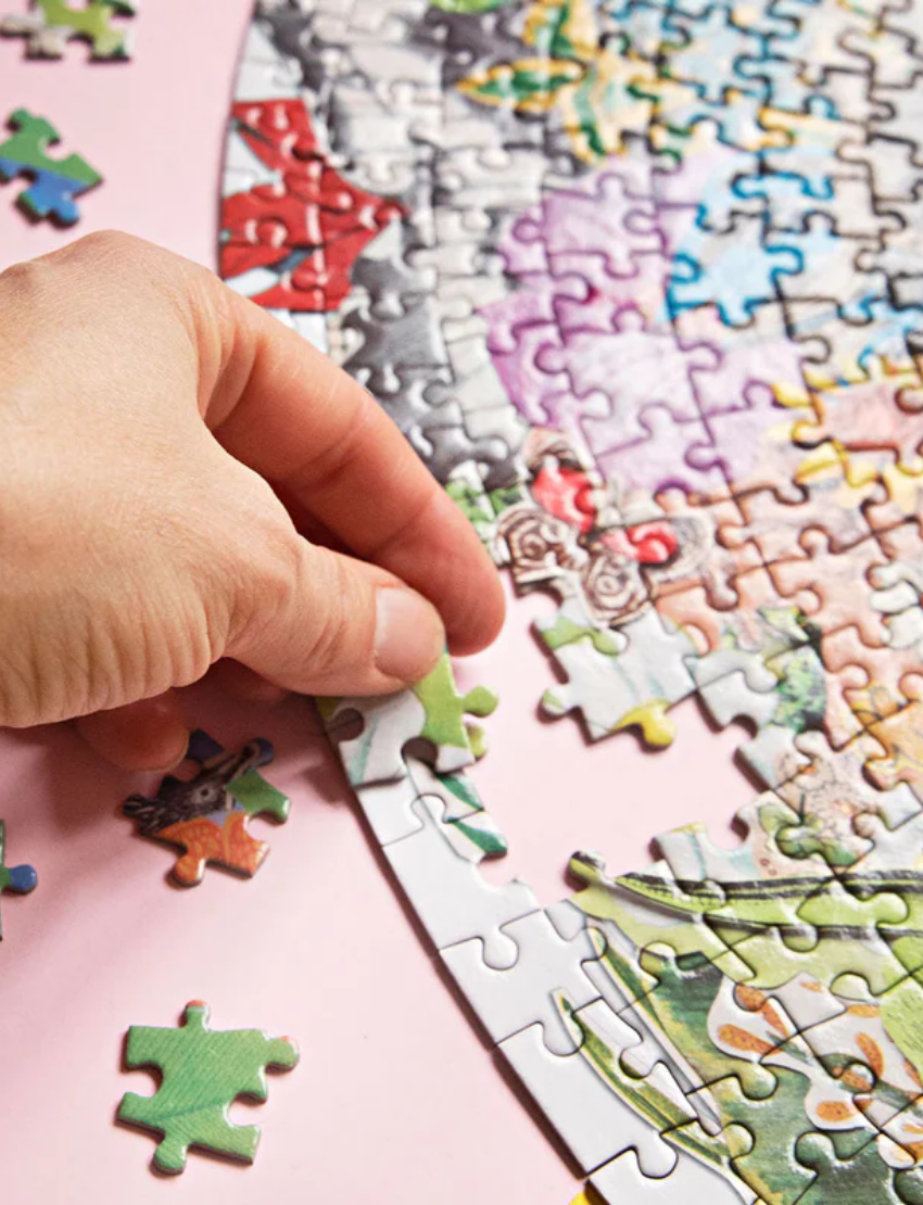 A round, colorful jigsaw puzzle with completed sections and scattered pieces is shown next to an open cylindrical container labeled "1000 Piece Puzzle" by Journey of Something. The puzzle features a vibrant design with various small images and shapes.