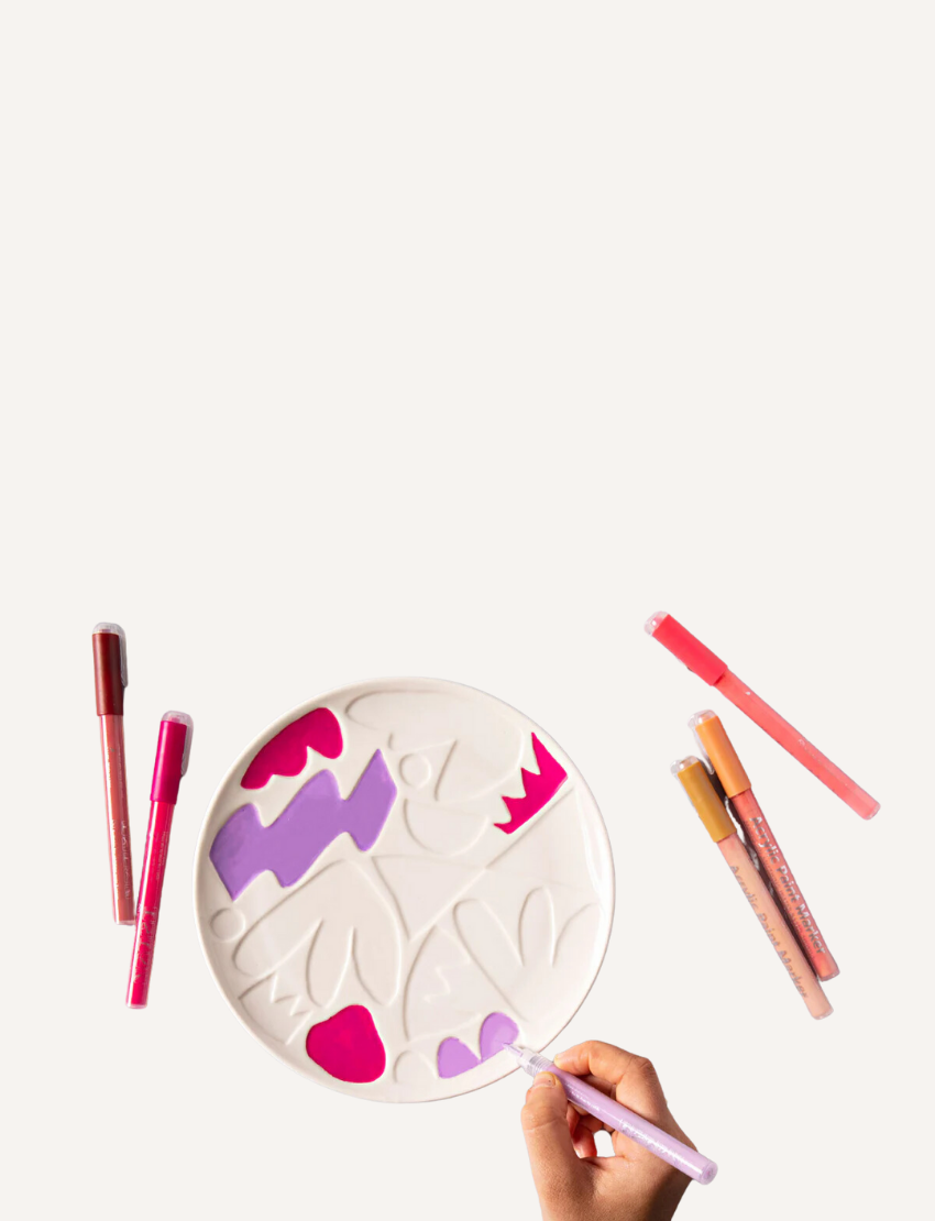 Image of the "Paint Your Own Plate - J.O.S Studio" by Journey of Something. The kit includes a plain ceramic plate with embossed leaf patterns, a set of colored markers, and a small palette. The packaging displays a partially colored plate as an example of the potential outcome.