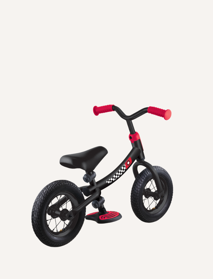 The Go Bike Air Balance Bike by Globber is a small, black balance bike featuring red handlebars and seat adjustments, specifically designed for toddlers aged 3-6. It includes two thick black tires and a checkered pattern on the frame near the seat. The bike has no pedals, aiding young children in learning balance and coordination while developing essential motor skills.