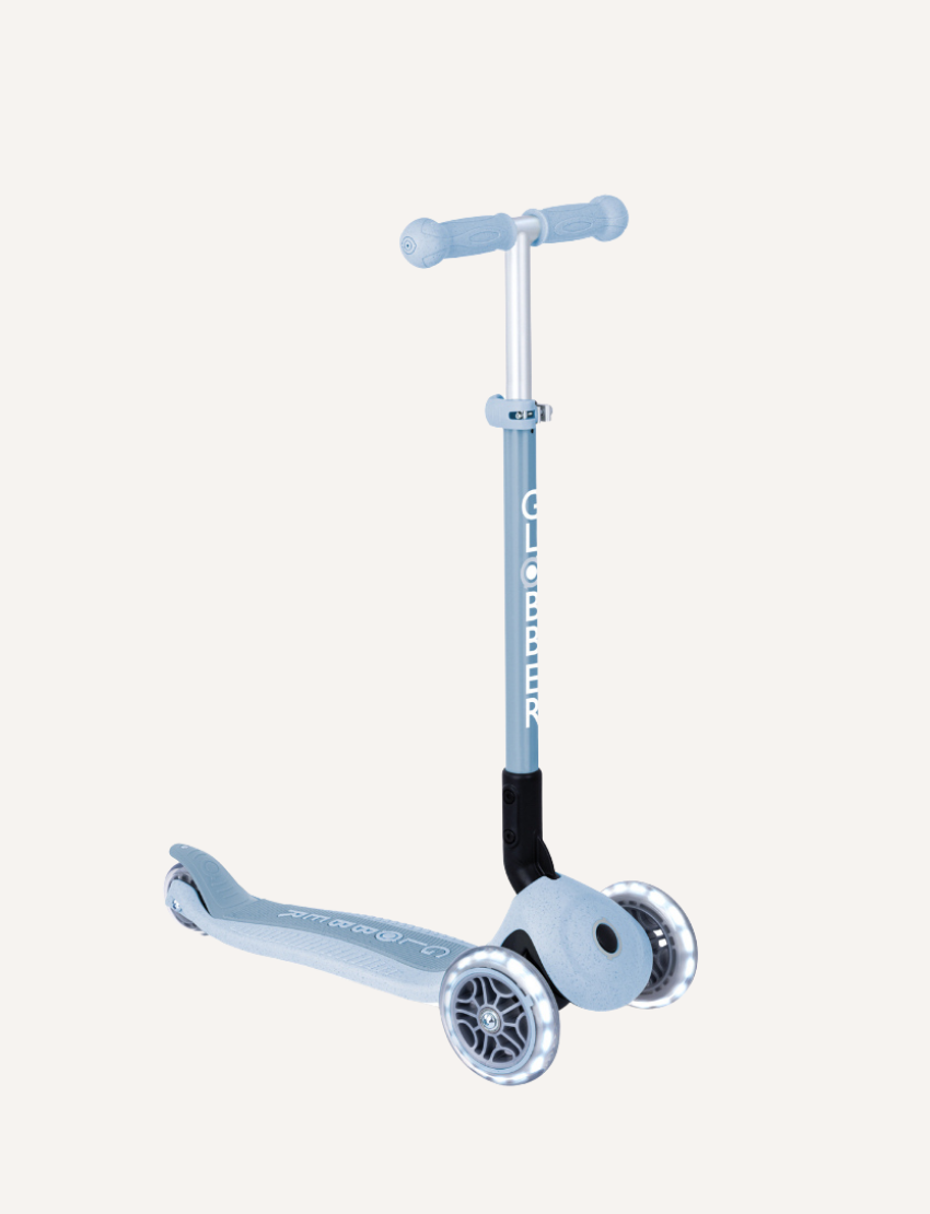 The Globber Ecologic Primo Foldable Scooter with Lights in light blue has LED front wheels, a three-wheel design, and an adjustable aluminium T-bar. The deck displays "GLOBBER," and larger front wheels provide a sleek design suited for young riders.