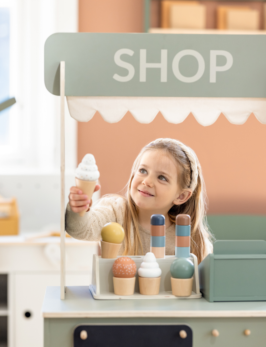 The Flexa Shop and Cafe is a grey play shop adorned with a "SHOP" sign above a charming scalloped awning. It features a counter, shelf, and knobs, measures 113.6 cm in height, 60 cm in width, 36.4 cm in depth, and stands on four short cylindrical legs.