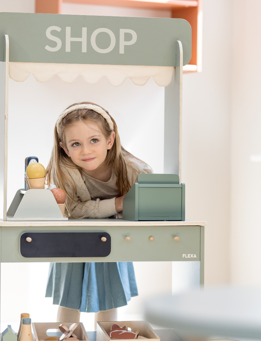 The Flexa Shop and Cafe is a grey play shop adorned with a "SHOP" sign above a charming scalloped awning. It features a counter, shelf, and knobs, measures 113.6 cm in height, 60 cm in width, 36.4 cm in depth, and stands on four short cylindrical legs.