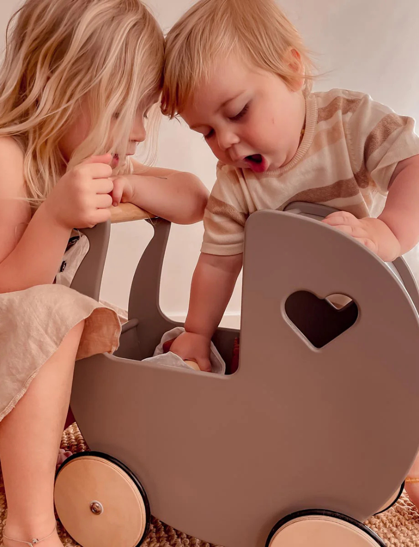 Two young children are playing with a Moover Classic Dolls Pram in off white. One child looks on while the other reaches inside. The pram features a heart-shaped cutout and large wooden wheels, and they play on a woven rug.