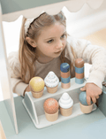 A young girl with long hair, wearing a headband and a light-colored sweater, is thoroughly enjoying her Flexa Ice Cream Set. The eco-friendly set includes various colorful, wooden ice cream cones arranged on a white shelf, fostering imaginative play as she reaches for one with delight.