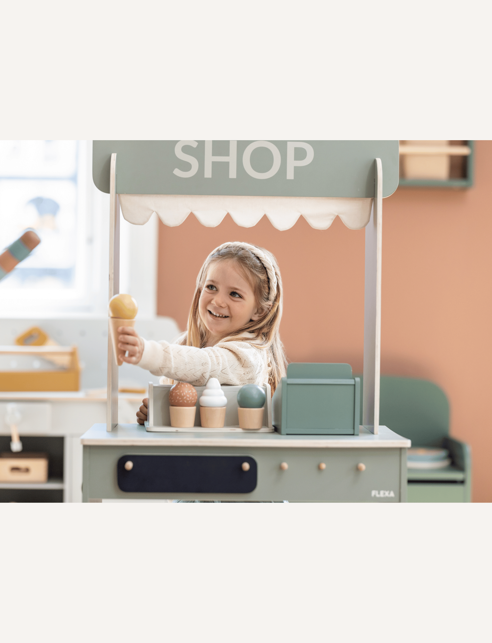 A young girl with long hair, wearing a headband and a light-colored sweater, is thoroughly enjoying her Flexa Ice Cream Set. The eco-friendly set includes various colorful, wooden ice cream cones arranged on a white shelf, fostering imaginative play as she reaches for one with delight.