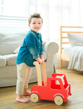 The Moover Push Truck is a red wooden toy car featuring beige wheels and handlebars. It boasts a simplistic design with cut-out windows and a small basket area at the back, marked with the word 